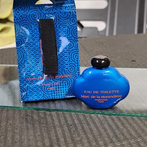 Pu&ograve; includere: Un flacone di profumo Eau de Toilette blu con un tappo nero accanto a una scatola blu con un cinturino nero. La bottiglia e la scatola hanno testo bianco. La bottiglia ha la forma di una nuvola.