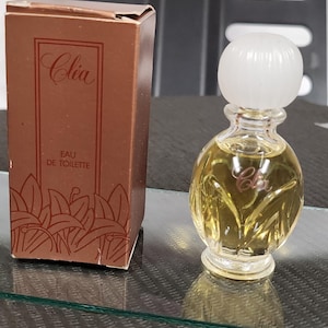 May include: A small, round perfume bottle with a white cap and a brown box. The bottle contains a yellow liquid and has a stylized "C" on the front. The box is brown with the name "Cléa" and "Eau de Toilette" printed on it.