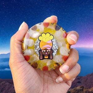 May include: A round, yellow resin art piece with embedded stones and a central graphic of a figure with a turban. The graphic is surrounded by silver glitter. The piece is held in a hand.