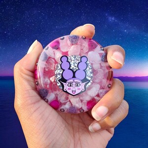 May include: A round, clear resin object filled with pink and white stones, and a cartoon face with purple hair. The object is held in a hand against a starry sky background. The cartoon face has a silver glitter background.