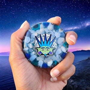 May include: A round, decorative object with a blue and white stone design, held in a hand. The center features a green and blue cartoon character. The background shows a starry night sky over a body of water.