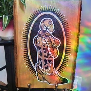 May include: Wooden art piece featuring a brown snake coiled around a cowboy boot with heart cutouts. The artwork is set within an oval frame with a sunburst design, all on a natural wood background. The piece has a rustic, country aesthetic.
