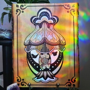 May include: A hand-painted wooden art piece featuring a stylized lamp design with butterflies and floral elements. The artwork is framed with decorative corner accents and a natural wood edge. The color palette includes black, white, and shades of brown and yellow.