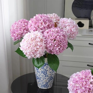 Pode incluir: Um buquê de flores de hortênsia rosa e roxa clara em um vaso com estampa azul e branca. As flores têm folhas verdes e são dispostas para criar uma exibição completa e exuberante. O vaso está em uma superfície preta.