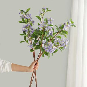 Könnte beinhalten: Ein Bund künstlicher Lavendelblüten mit grünen Blättern und braunen Stielen. Die Lavendelblüten sind hellviolett und die Blätter leuchtend grün. Die Blumen werden in der Hand einer Person gehalten.