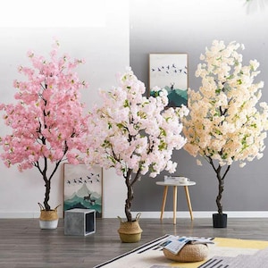 May include: Three artificial cherry blossom trees in shades of pink and cream. The trees have dark brown trunks and are potted in decorative baskets and a black pot. Artwork and a small table are in the background.