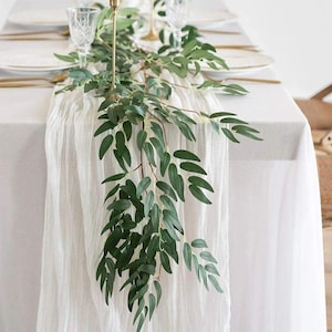 Puede incluir: Un mantel blanco está adornado con un camino de mesa blanco y transparente. Una guirnalda de hojas y ramas verdes adorna la mesa, creando un centro de mesa natural y elegante. La cubertería y los candelabros dorados añaden un toque de sofisticación.