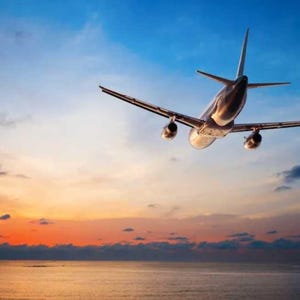 May include: A commercial airplane in flight, viewed from below, against a backdrop of a vibrant sunset over the ocean. The aircraft is silver and black, with its wings and engines clearly visible. The sky transitions from blue to orange and yellow.