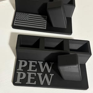 Può includere: Due organizer da scrivania neri con tre scomparti ciascuno. Uno ha una bandiera americana grigia, l'altro le parole "PEW PEW" in grigio. Ogni organizer ha un piccolo blocco angolato.