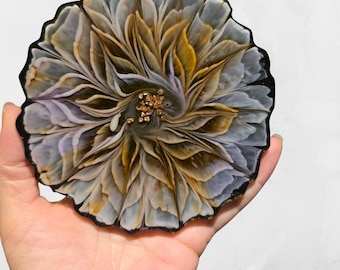 May include: A resin coaster with a floral design. The coaster has a black edge with gold flecks, and a central flower design in white, gold, and purple. The coaster is round and has a glossy finish.