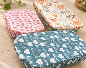 May include: Three rectangular fabric bowl covers with elastic edges. One cover features a pink strawberry pattern, one has a sun and leaf motif in orange and brown, and the third is dark teal with white hearts and the text live, love, smile, laugh.