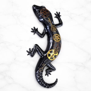 May include: A black lizard sculpture with bronze and gold-colored gears and keys. The steampunk-style reptile is posed on a white marble surface. The lizard's body is detailed with mechanical elements.