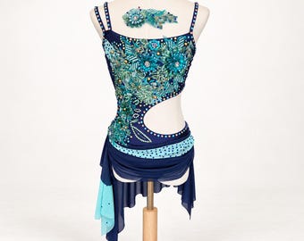 May include: A dark blue dance dress featuring an asymmetrical cutout at the waist, adorned with intricate teal and green floral beadwork and sequins. The skirt includes layered chiffon panels in dark blue and light blue, with a matching hair accessory.