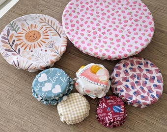 May include: A set of seven round fabric bowl covers in various sizes and patterns, including pink strawberries, a sun and foliage print, blue hearts with the word love, beige gingham, and red, white, and blue American flag designs.