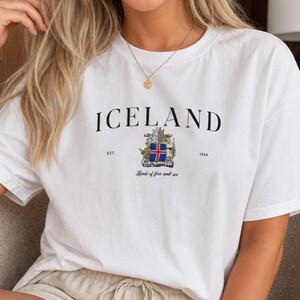 Puede incluir: Camiseta blanca con la palabra "ICELAND" en negro, un escudo y el texto "Est. 1944". La camiseta se combina con pantalones cortos beige. La camiseta tiene cuello redondo y mangas cortas.