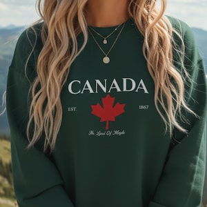 Könnte beinhalten: Forstgrünes Sweatshirt mit dem Wort "CANADA" in Weiß, einem roten Ahornblatt und dem Text "The Land Of Maple". Das Sweatshirt hat einen Rundhalsausschnitt.