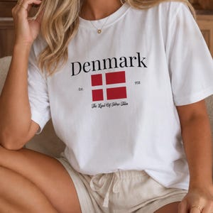 Könnte beinhalten: Ein weißes kurzärmeliges T-Shirt mit dem Wort Denmark in schwarzer Schrift über einer rot-weißen dänischen Flagge. Unter der Flagge steht der Text Est. 958 und The Land Of Fairy Tales. Ein kleines kreisförmiges Logo oben rechts zeigt Artwork by Soft Groove Design.