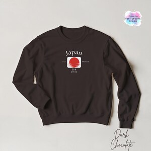 May include: Dark chocolate-colored sweatshirt with a graphic featuring the word "Japan" above a red sun design. The design includes Japanese characters and the text "EST 660BCE". The sweatshirt is a crew neck style.