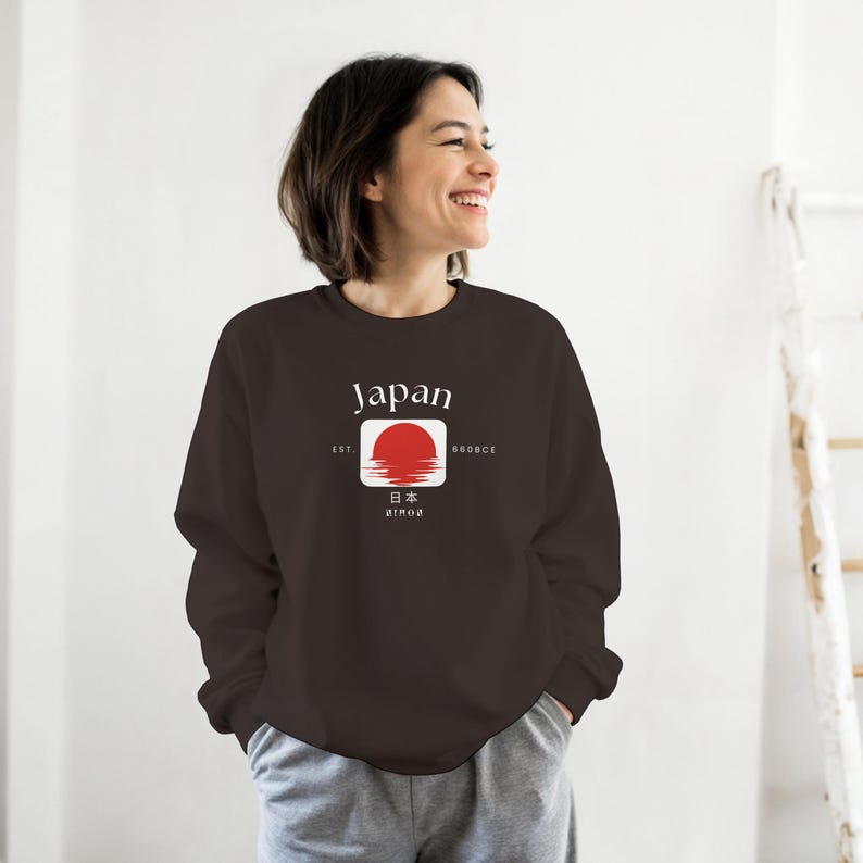 May include: A dark brown sweatshirt with a white square graphic featuring a red sun and the word "Japan" above it. The graphic also includes Japanese characters and the text "Est 660BCE". The sweatshirt is worn by a person.