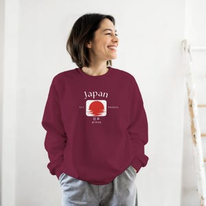 May include: Burgundy sweatshirt with a white and red graphic featuring the word "Japan" above a red sun design. The sweatshirt has a crew neck and long sleeves. The text "EST. 600BCE" and Japanese characters are below the sun graphic.