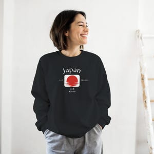 May include: Black sweatshirt with the word "Japan" in white above a red sun design inside a white square. The sweatshirt also has Japanese text and the words "EST" and "660BCE". The person is wearing gray sweatpants.