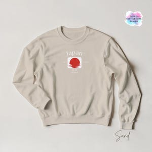 May include: Sand-colored sweatshirt with a crew neck. The front features a graphic of a red sun over water within a white square, with the word "Japan" above. The sweatshirt also has the text "EST. since" and "PRODUCT" on the front.