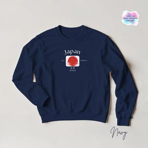 May include: Navy blue sweatshirt featuring a graphic design with the word "Japan" in white, a red sun, and Japanese text. The sweatshirt has a crew neck and long sleeves. The design includes the text "EST 660BCE".