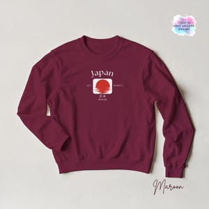 May include: Maroon crewneck sweatshirt with a white and red graphic featuring the word "Japan" and a rising sun design. The sweatshirt also includes Japanese characters and the text "EST 660BCE".