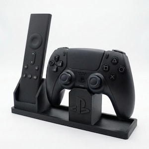 May include: A black, 3D-printed stand holding a remote control and a game controller. The stand has a rectangular base with a slot for the remote and a raised platform with the PlayStation logo for the controller.