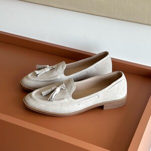 May include: A pair of light beige suede loafers with tassel detailing. The shoes feature a classic slip-on design with a low heel and a rounded toe. The loafers are displayed on a brown surface.