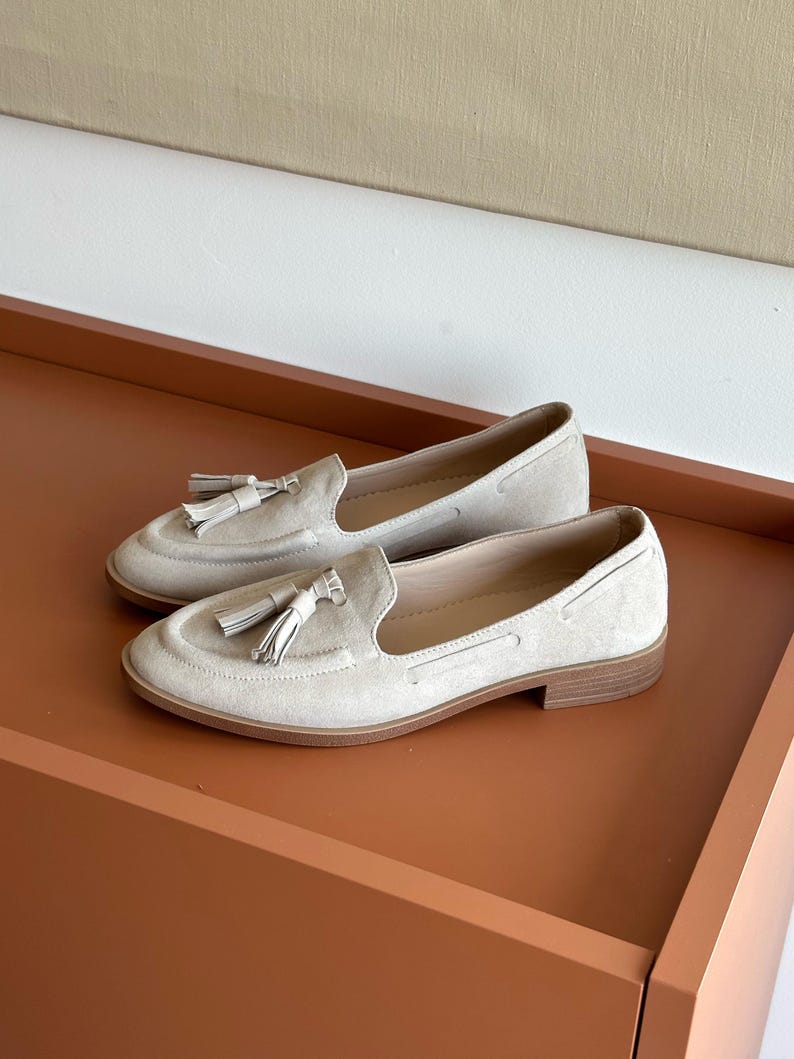 May include: A pair of light beige suede loafers with tassel detailing. The shoes feature a classic slip-on design with a low heel and a rounded toe. The loafers are displayed on a brown surface.