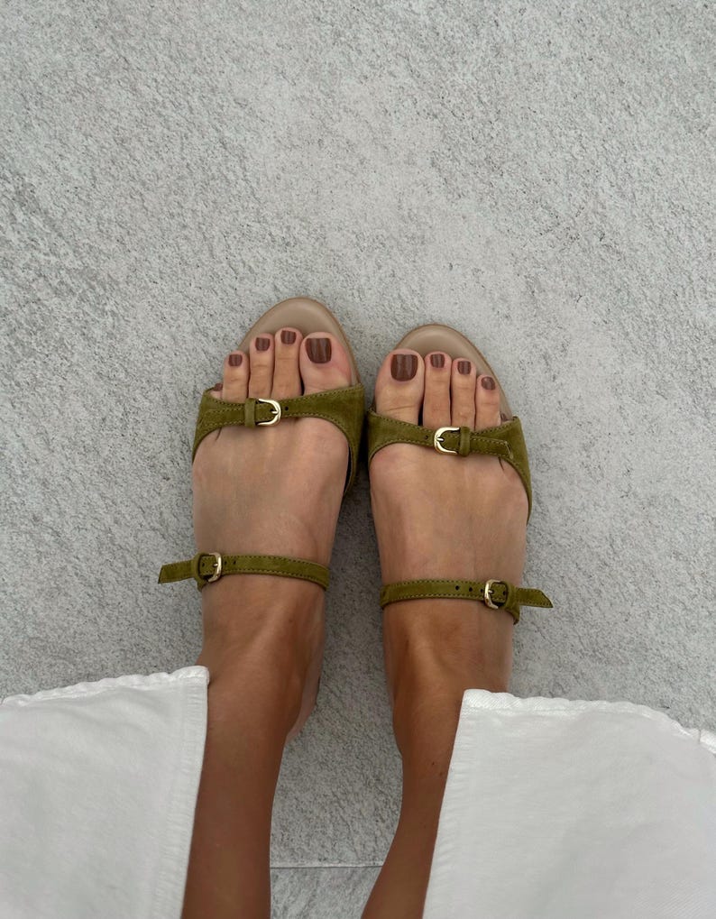 May include: Olive green sandals with ankle straps and small gold buckles. The sandals have a toe strap and a small buckle detail on the top of the foot. The background is a light gray surface.