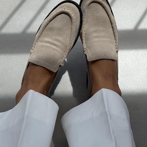 May include: A pair of beige suede loafers with black soles. The shoes are worn with white pants. The loafers have a classic slip-on design and are made of a soft, textured material. The image is taken in natural light.