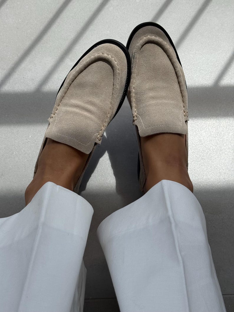 May include: A pair of beige suede loafers with black soles. The shoes are worn with white pants. The loafers have a classic slip-on design and are made of a soft, textured material. The image is taken in natural light.