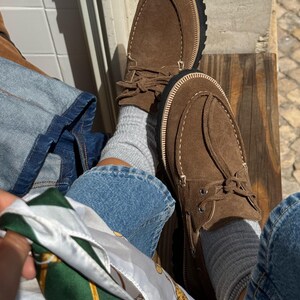 May include: Brown suede moccasin-style shoes with a thick black sole and cream-colored stitching. The shoes have a rounded toe and a leather tie detail. The image also shows blue jeans, gray socks, and a patterned scarf.
