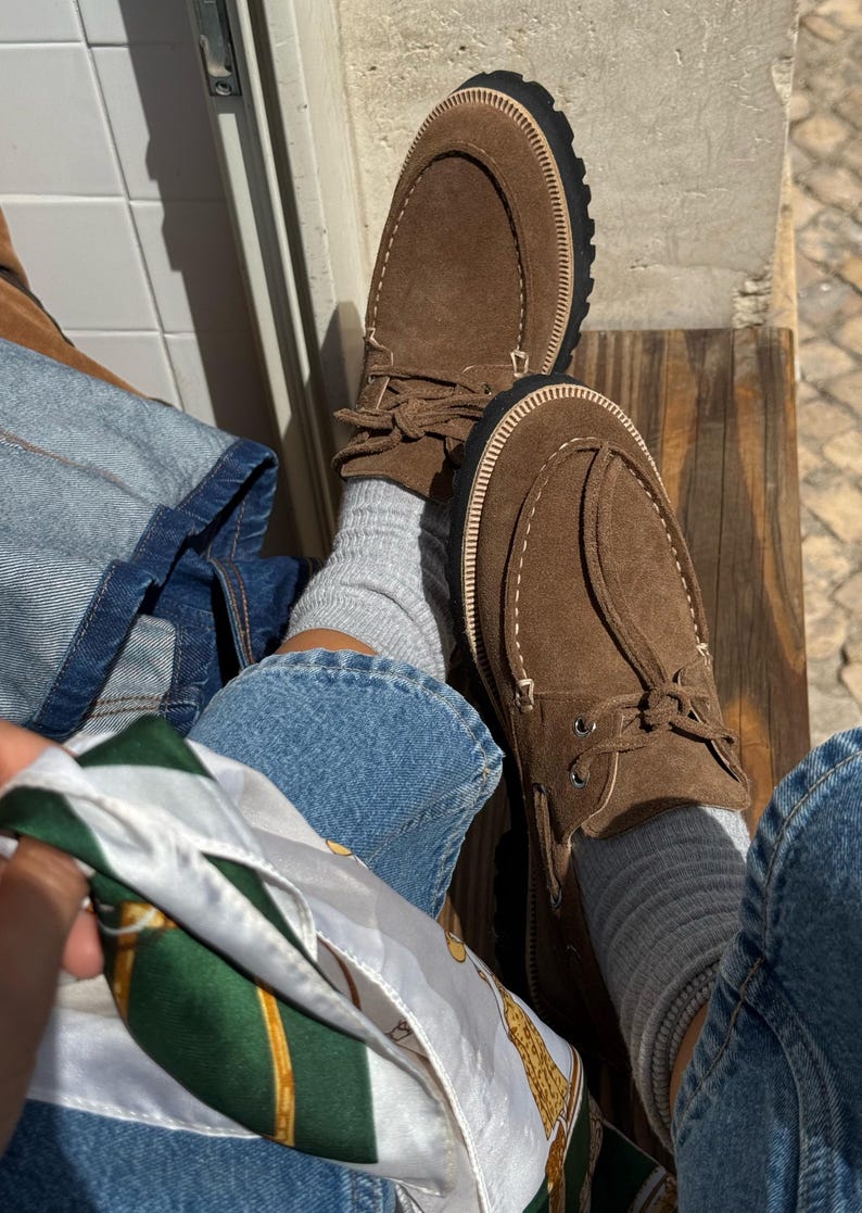 May include: Brown suede moccasin-style shoes with a thick black sole and cream-colored stitching. The shoes have a rounded toe and a leather tie detail. The image also shows blue jeans, gray socks, and a patterned scarf.