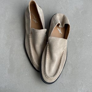 May include: A pair of beige suede loafers with a rounded toe and a slip-on design. The shoes have a black sole and a stitched seam around the upper. The interior of the shoe is a lighter beige color with the word "Aspen" printed in white.