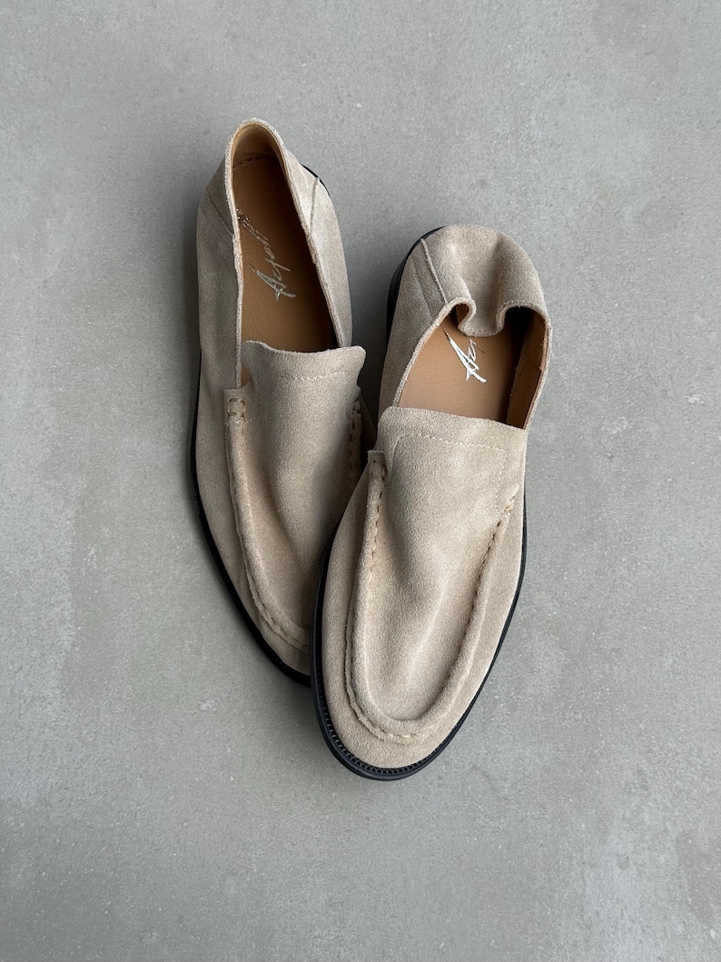 May include: A pair of beige suede loafers with a rounded toe and a slip-on design. The shoes have a black sole and a stitched seam around the upper. The interior of the shoe is a lighter beige color with the word "Aspen" printed in white.