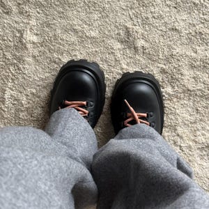 May include: A pair of black leather boots with thick, ridged soles and pink laces. The boots are worn with gray sweatpants, creating a casual, everyday look. The boots have a rugged, outdoor style.