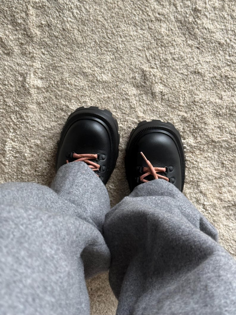 May include: A pair of black leather boots with thick, ridged soles and pink laces. The boots are worn with gray sweatpants, creating a casual, everyday look. The boots have a rugged, outdoor style.