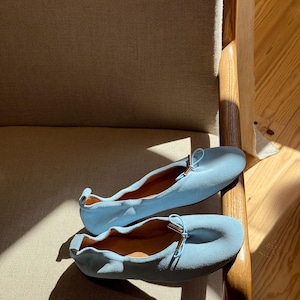 May include: A pair of light blue ballet pumps featuring a soft, matte texture, a rounded toe, and a small decorative bow at the front with metallic gold-toned hardware accents. The shoes have a gathered, elasticised topline and a pull tab at the heel.