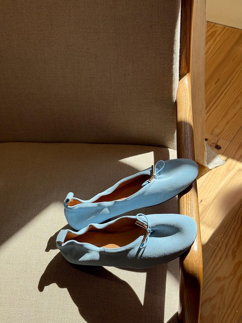 May include: A pair of light blue ballet pumps featuring a soft, matte texture, a rounded toe, and a small decorative bow at the front with metallic gold-toned hardware accents. The shoes have a gathered, elasticised topline and a pull tab at the heel.