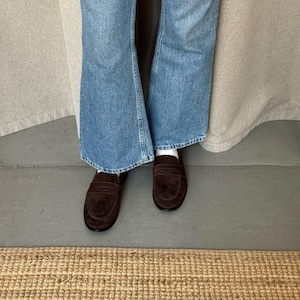 May include: A pair of dark brown suede penny loafers. The shoes are worn with blue denim jeans. The jeans have a flared leg opening. The shoes are on a gray surface with a woven rug in the foreground.