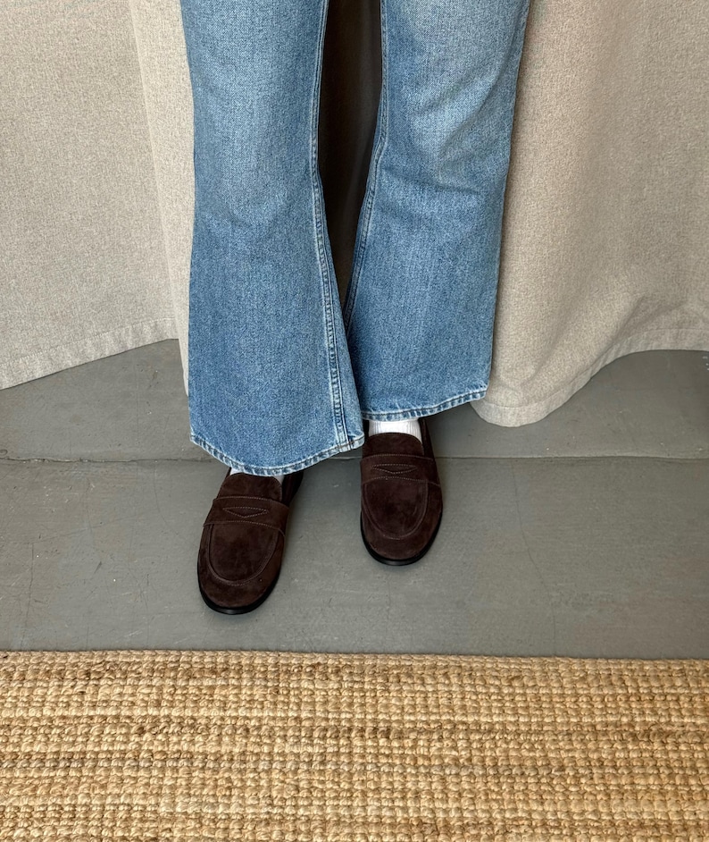 May include: A pair of dark brown suede penny loafers. The shoes are worn with blue denim jeans. The jeans have a flared leg opening. The shoes are on a gray surface with a woven rug in the foreground.
