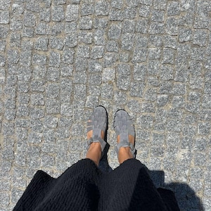 Puede incluir: Vista superior de sandalias grises con correas tejidas. Las sandalias se usan con una falda negra, sobre una superficie adoquinada. Las sandalias tienen un cierre de hebilla.