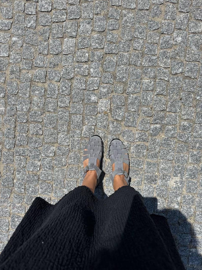 Puede incluir: Vista superior de sandalias grises con correas tejidas. Las sandalias se usan con una falda negra, sobre una superficie adoquinada. Las sandalias tienen un cierre de hebilla.