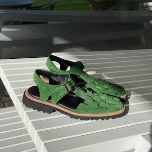Puede incluir: Sandalias de ante verde con dise&ntilde;o trenzado en la parte superior, suela negra y ribete marr&oacute;n. Las sandalias tienen cierre de hebilla. Se muestran sobre una mesa blanca con lamas. Miden unos 25 cm.