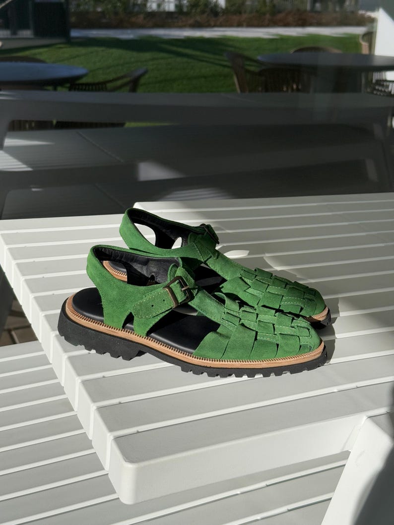Puede incluir: Sandalias de ante verde con dise&ntilde;o trenzado en la parte superior, suela negra y ribete marr&oacute;n. Las sandalias tienen cierre de hebilla. Se muestran sobre una mesa blanca con lamas. Miden unos 25 cm.