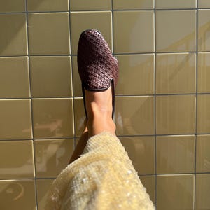 May include: A woven brown leather loafer with a black trim. The shoe is shown against a background of yellow tiles. The shoe is a slip-on style.
