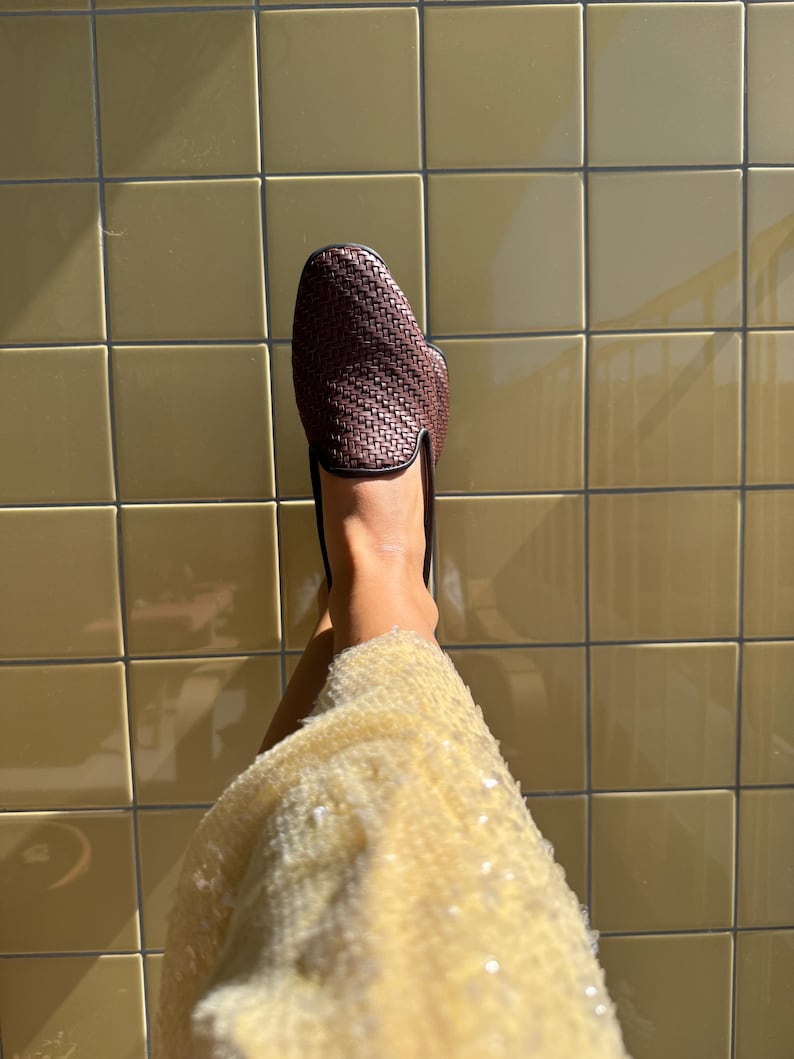 May include: A woven brown leather loafer with a black trim. The shoe is shown against a background of yellow tiles. The shoe is a slip-on style.
